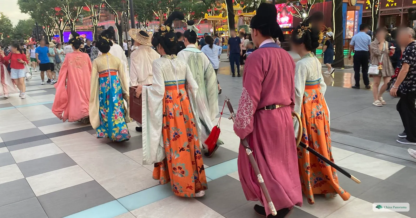 A group in Tang-style dress — long orange, blue and pink robes, one with a sword — walking away down Grand Tang Mall among evening crowds