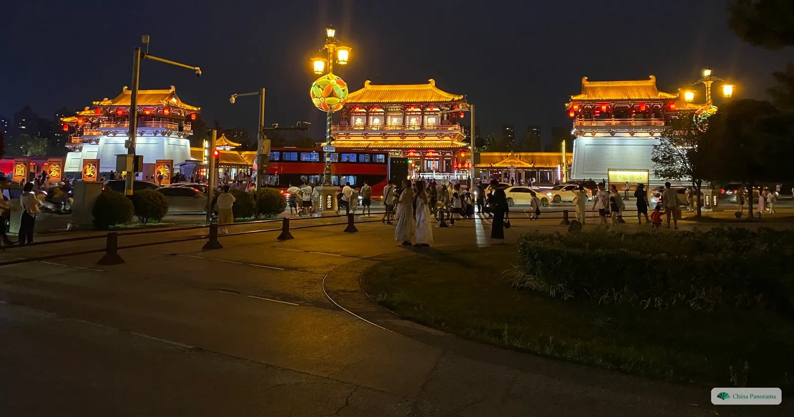 Three Tang-style pavilions outlined in golden light across a road at night, with pedestrians and a red bus passing in front