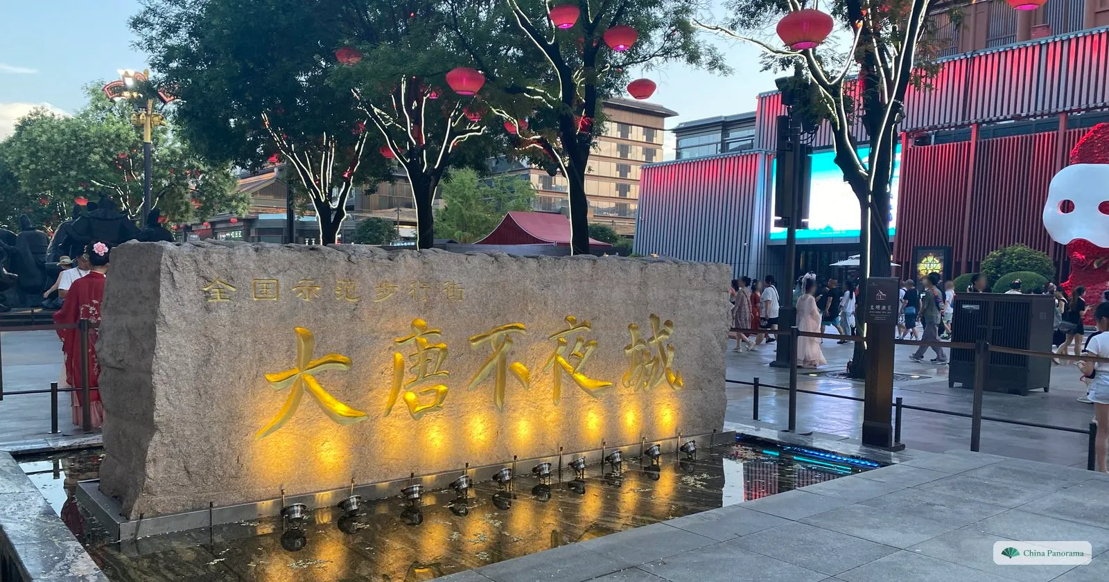 A rough block of stone carved with the gilded characters 大唐不夜城, lit from below at dusk, with visitors and red lanterns in the trees behind