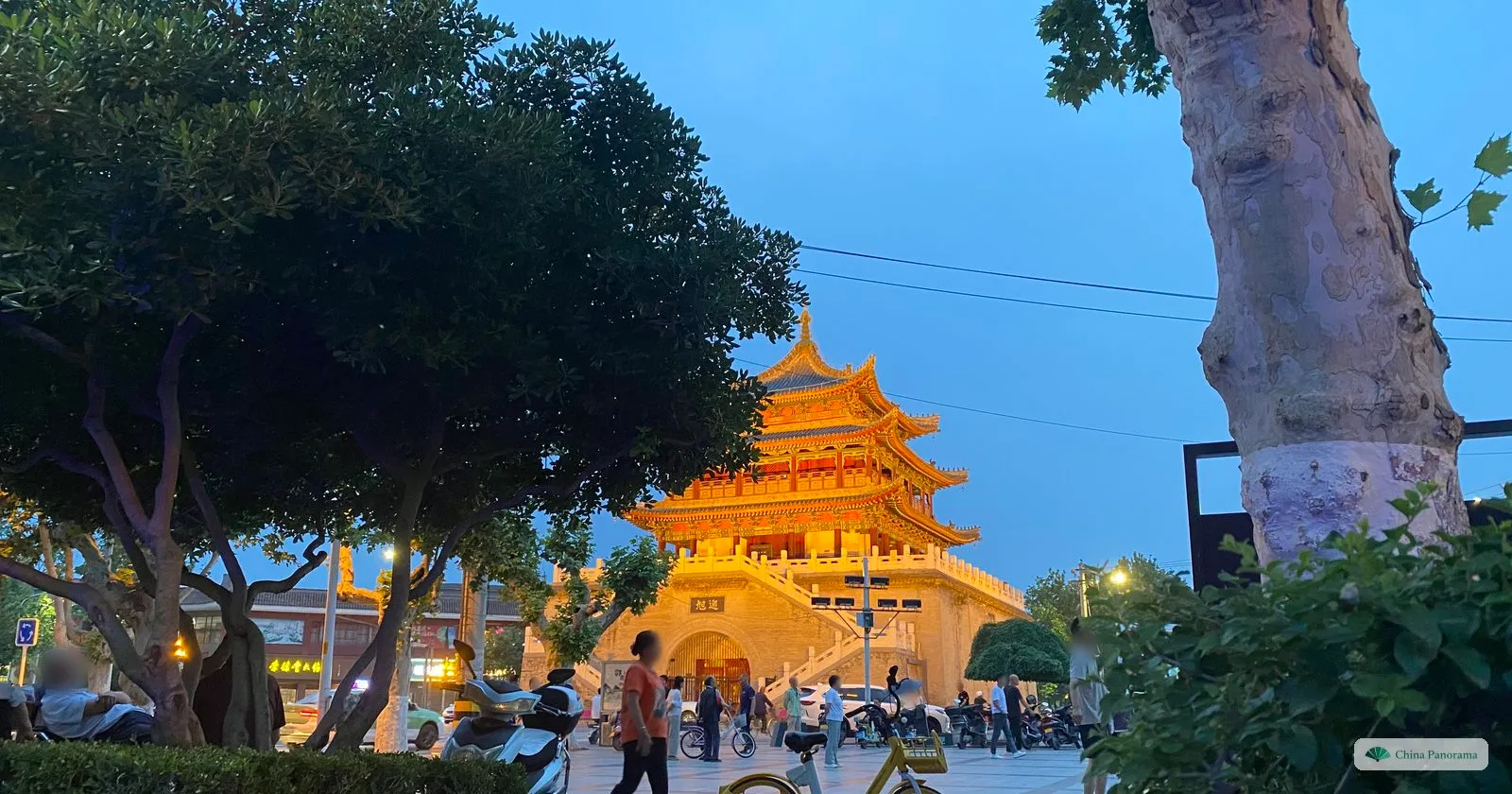 The Huyi Bell Tower floodlit in gold at dusk above a paved square, with the plaque 迎旭 over its gateway, locals sitting and walking under trees, and a plane tree and shrubs in the foreground