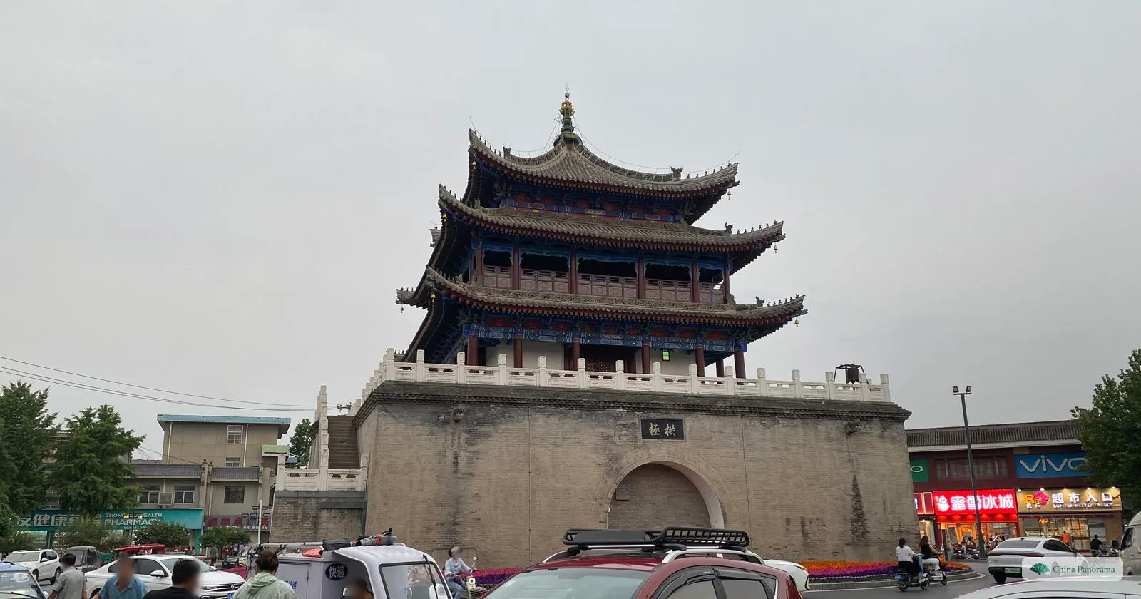 The Huyi Bell Tower, a three-eaved timber tower with red and blue painted beams on a brick base with a white balustrade and an arched gateway under the plaque 拱极, seen from the junction with cars and scooters in the foreground under a grey sky