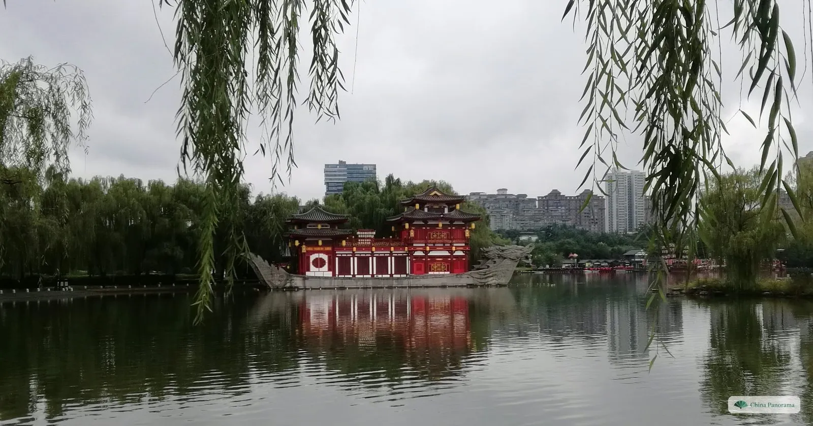 A red two-storey pavilion on a boat-shaped stone base at the edge of the lake in Tang Paradise, framed by willow branches