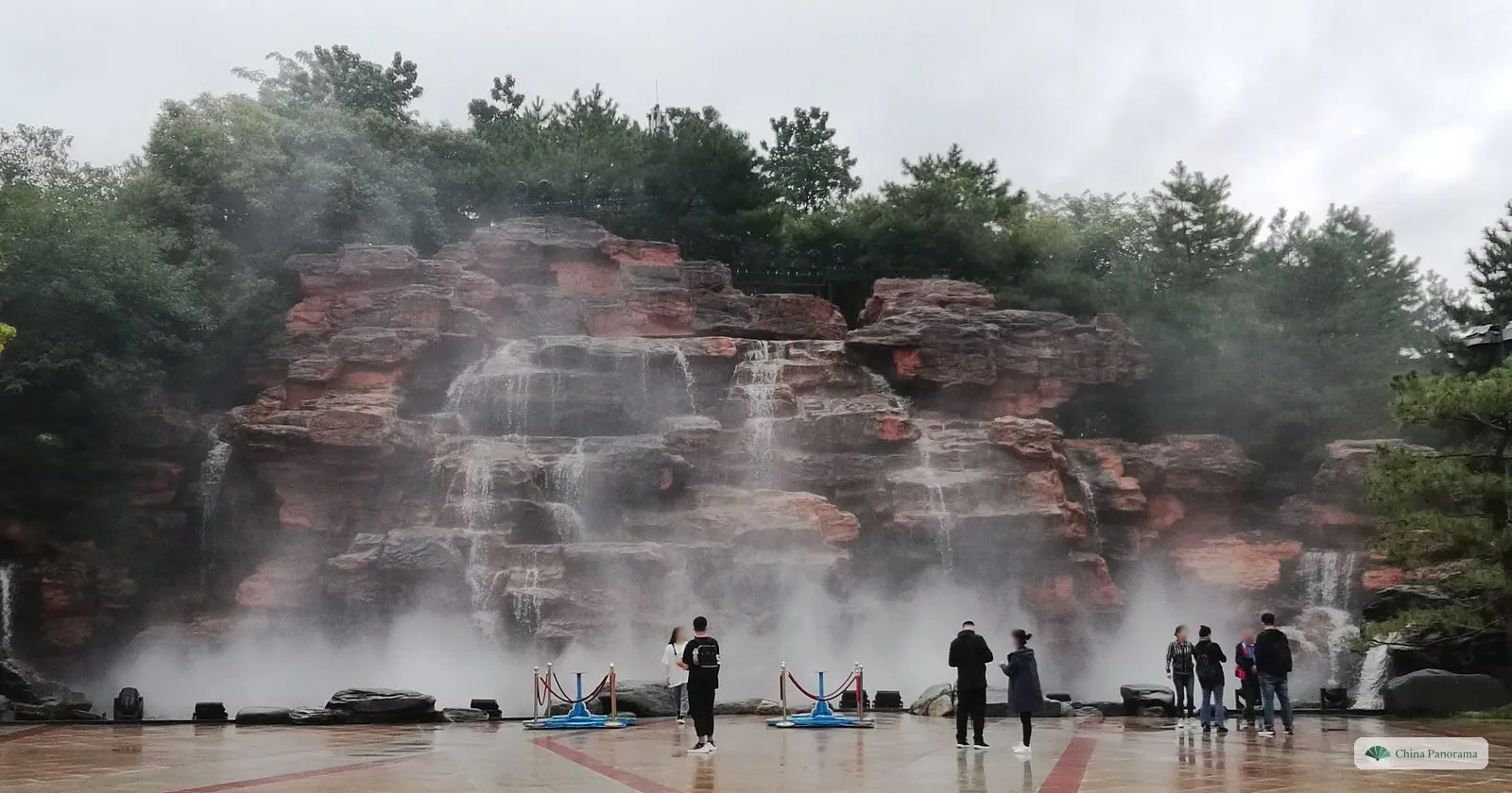 An artificial waterfall over red rock in Tang Paradise, wrapped in mist, with a few visitors standing in front of it