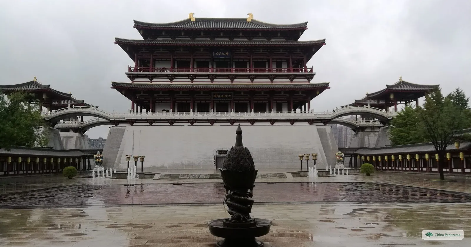 The Ziyun Tower in Tang Paradise, a tall red-and-white hall on a grey stone terrace with arched bridges to its side pavilions, reflected in rain-wet paving