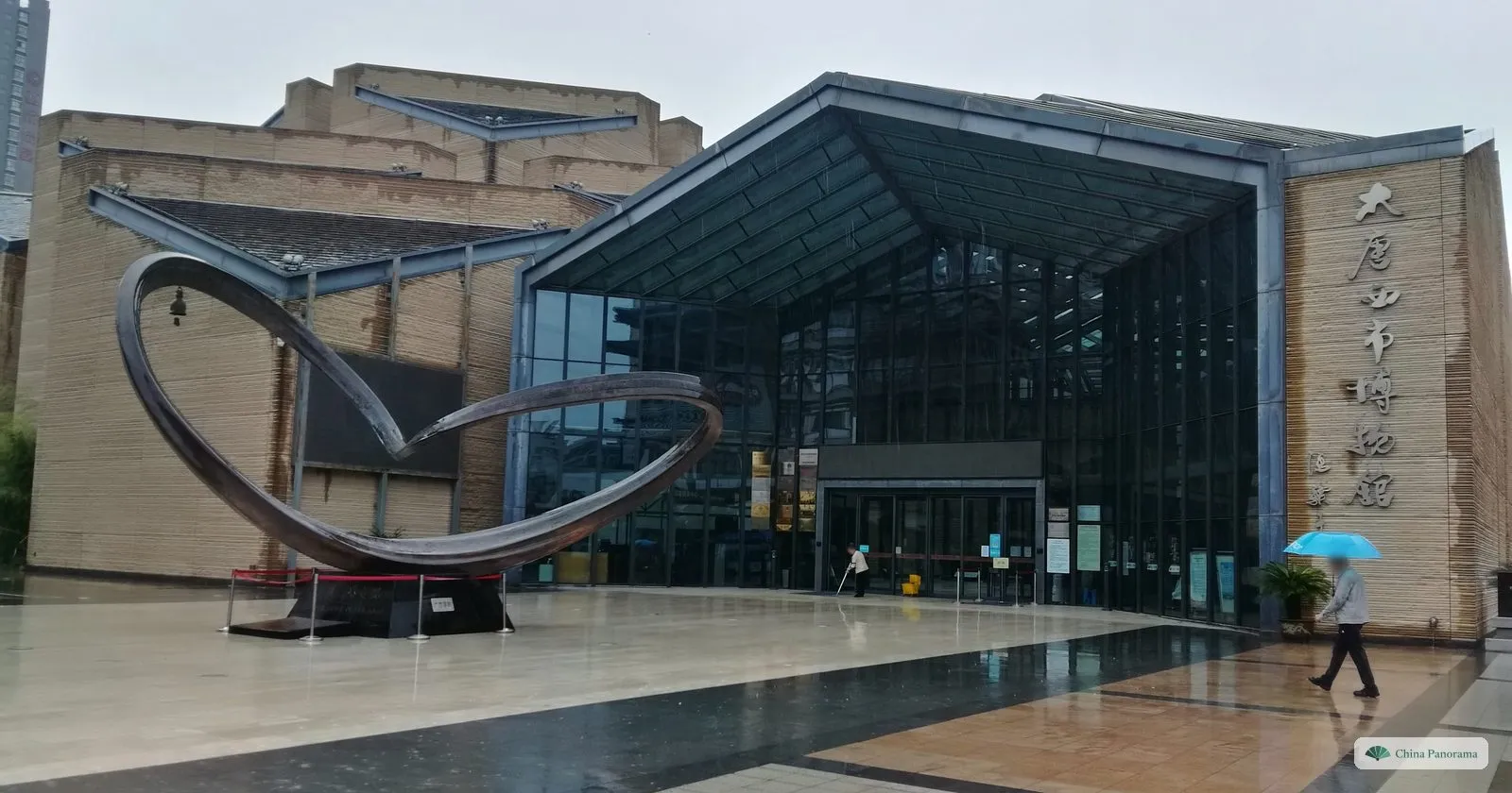The glass-fronted entrance of the Tang West Market Museum, with its name in metal characters on a stone wall and a large ring-shaped steel sculpture on the forecourt