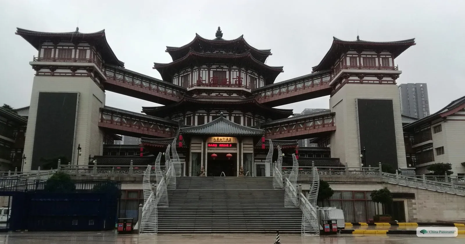 The main building of Tang West Market: a Tang-style hall with layered grey roofs, linked by raised walkways to two side towers, above a broad flight of steps in the rain
