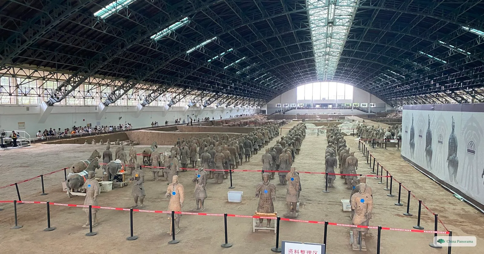 Reassembled terracotta soldiers and horses standing behind a rope barrier at the back of Pit 1, some with blue tags on their chests, under the steel arches of the hall