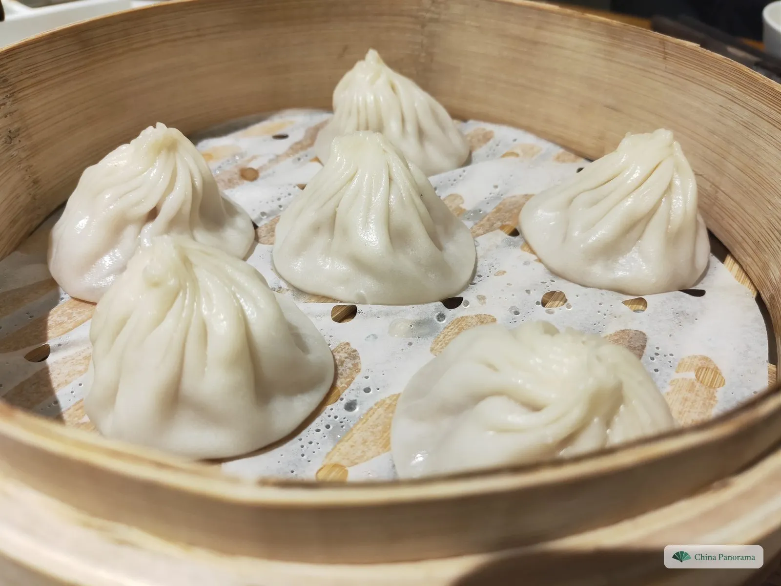 Six soup dumplings in a bamboo steamer lined with perforated paper, their thin white wrappers gathered into twisted knots on top
