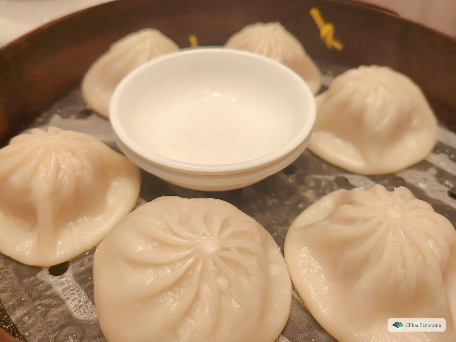 A steamer basket with a small white dish in the middle, surrounded by pleated soup dumplings
