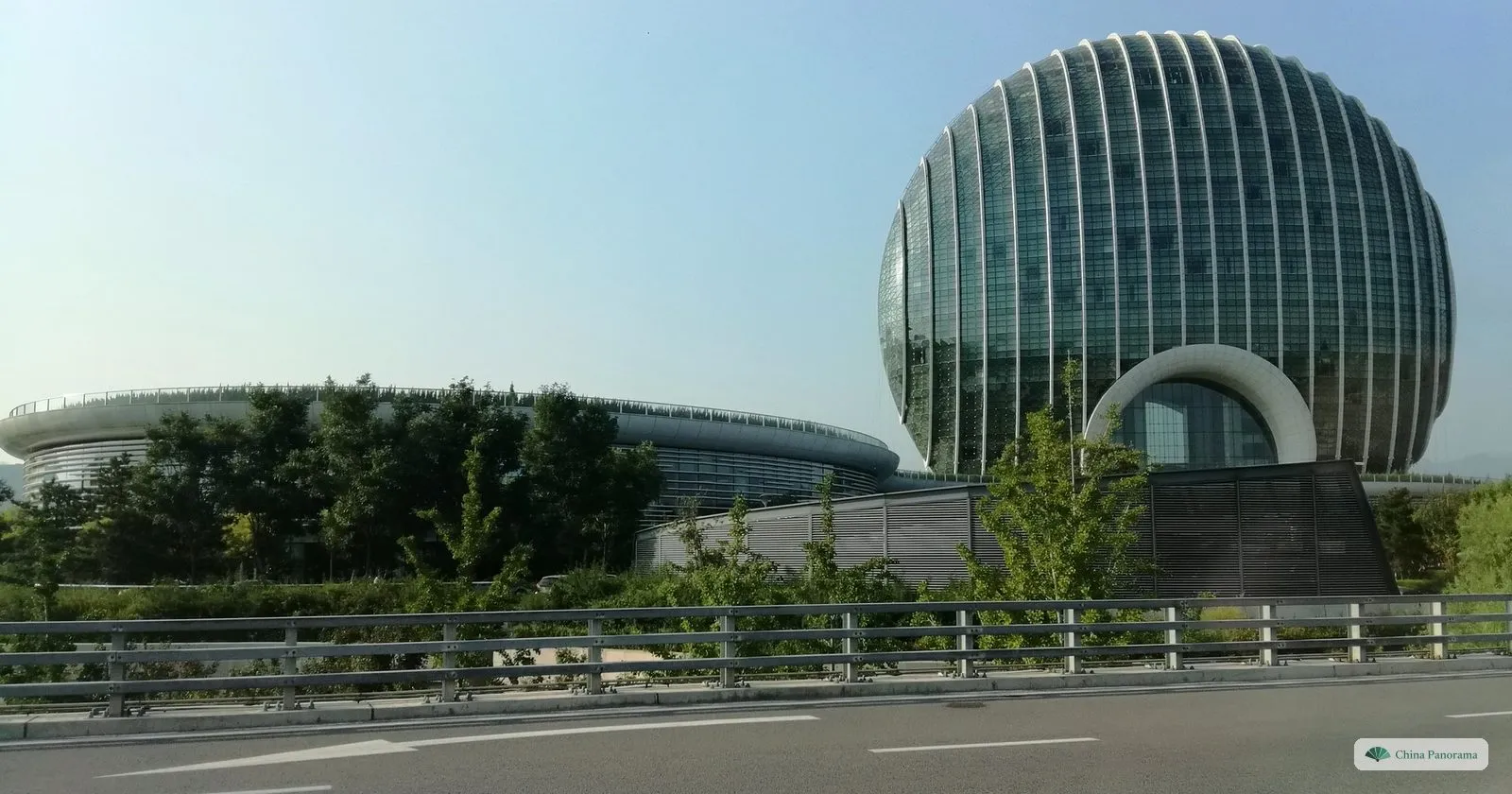 A hotel in the shape of a giant upright glass disc beside Yanqi Lake, seen from a road bridge