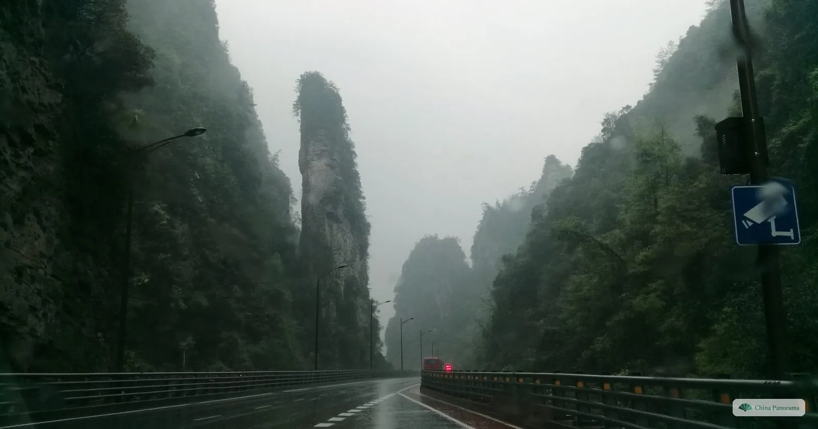 A tall, slender sandstone pillar with trees on its top rising beside a wet road in a gorge, with more pillars fading into low cloud behind it
