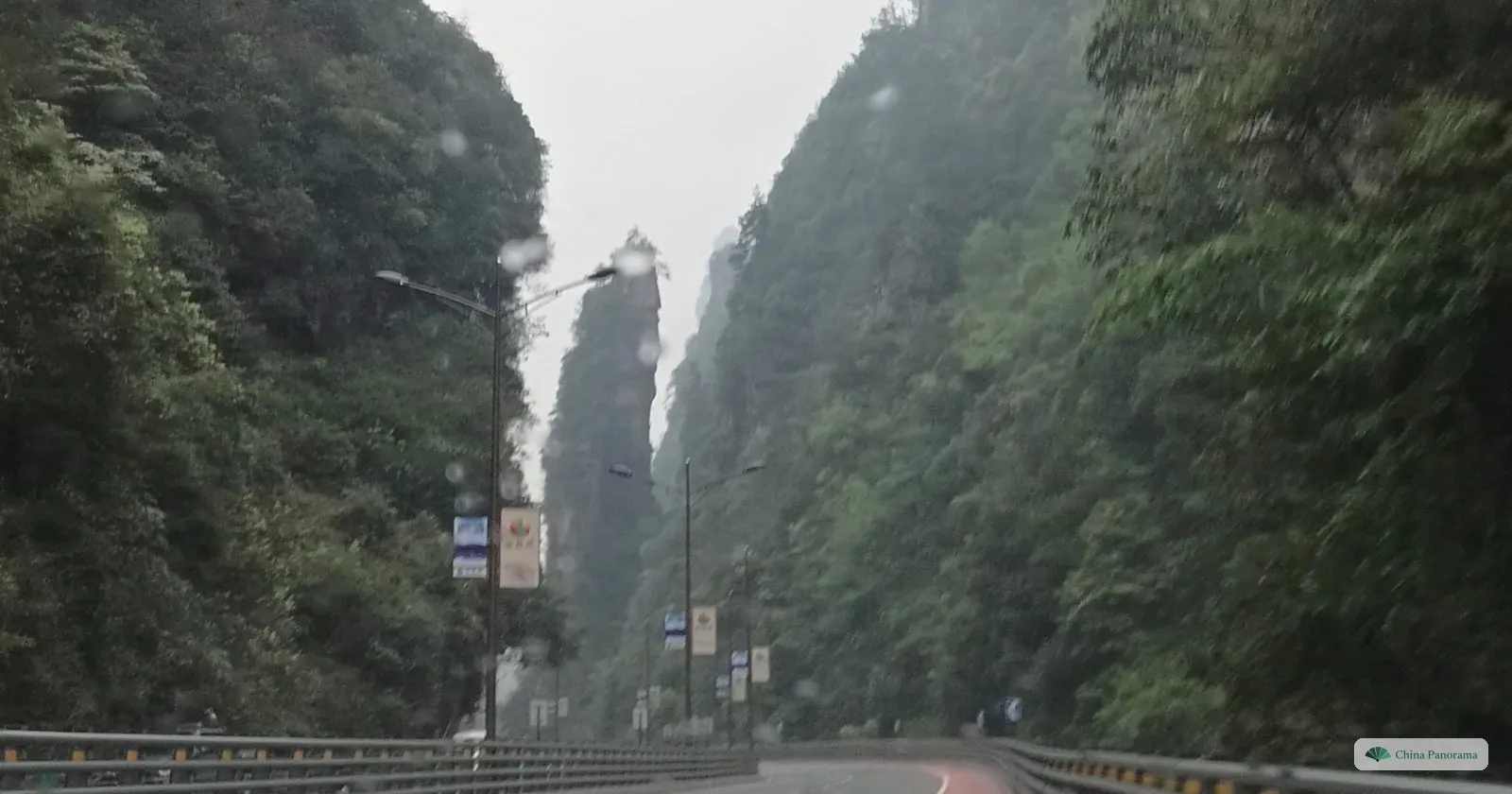 A road curving between forested cliffs, with a narrow rock pillar standing in the gap ahead and raindrops on the windscreen