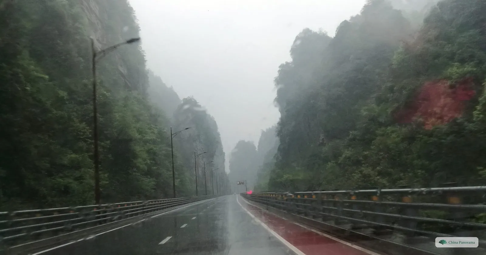 A straight wet road between steep wooded rock walls in heavy rain, cloud hanging in the gorge and a patch of red earth on the right-hand slope