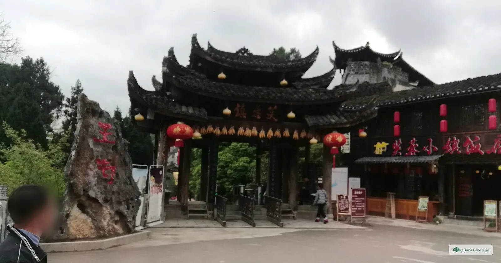 The wooden entrance gate of Furong Town with layered upswept roofs, a board reading 芙蓉镇, a row of dried corn cobs and red lanterns, a tall rock with red characters on the left