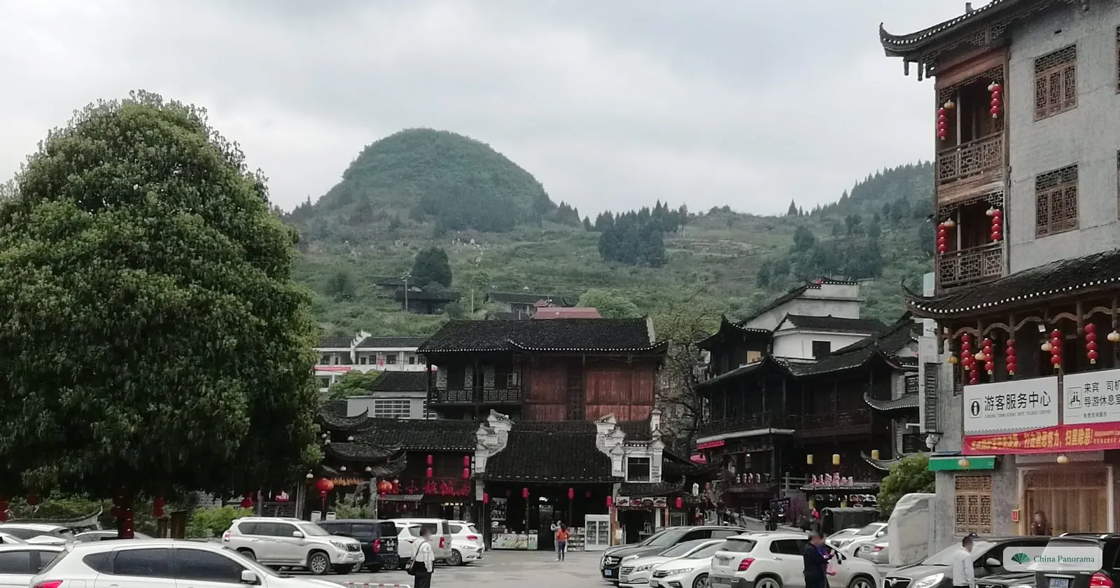 A parking square in Furong Town in front of dark timber buildings with red lanterns, a visitor centre on the right and a rounded green hill behind