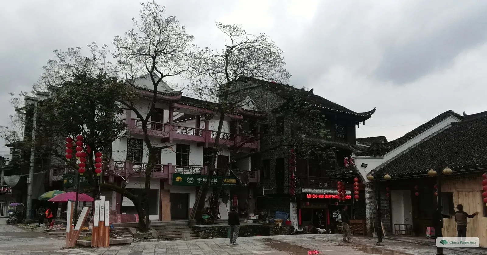 A paved square in Furong Town with a pink-and-white two-storey building with wooden balconies, bare trees, red lanterns and a few people