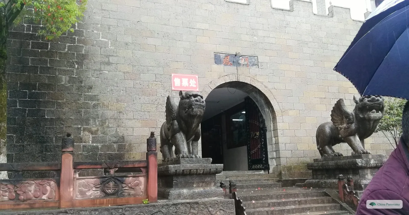 An arched gate in a high grey stone wall with crenellations, two winged stone lions on pedestals either side, a red sign reading 售票处 (ticket office) and an umbrella at the edge of the frame