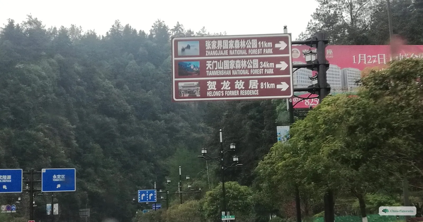 A brown tourist sign over a road with three bilingual lines — Zhangjiajie National Forest Park 11 km, Tianmenshan National Forest Park 34 km, He Long’s Former Residence 81 km — against wooded hills, with blue road signs lower left