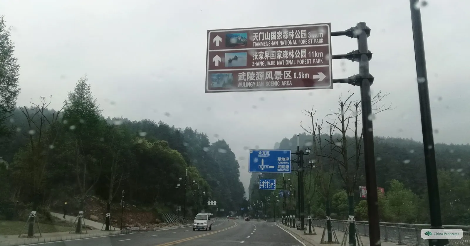 A wide road between wooded hills with a brown sign on a post listing Tianmenshan National Forest Park, Zhangjiajie National Forest Park 11 km and Wulingyuan Scenic Area 0.5 km