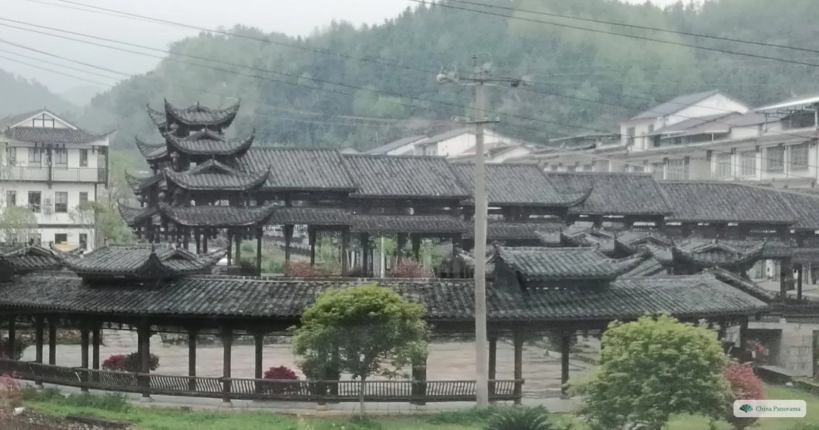 A long covered wooden bridge with grey tiled roofs and several tiered pavilion towers, white houses and green hills behind