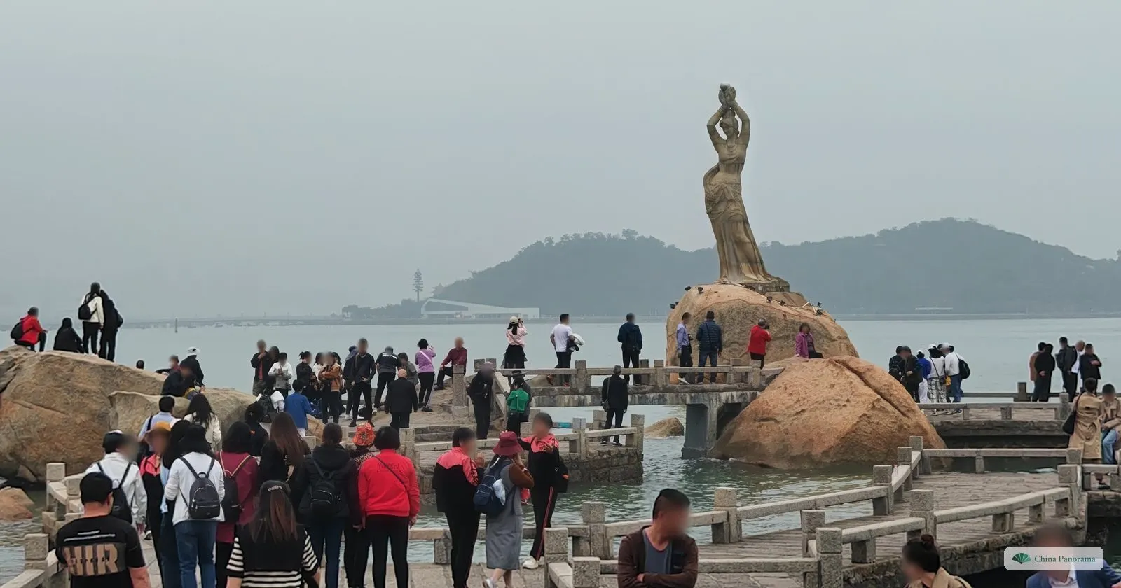 Visitors on the rocks and causeway in front of the granite Fisher Girl statue in Zhuhai, holding a pearl above her head, with a hilly island behind