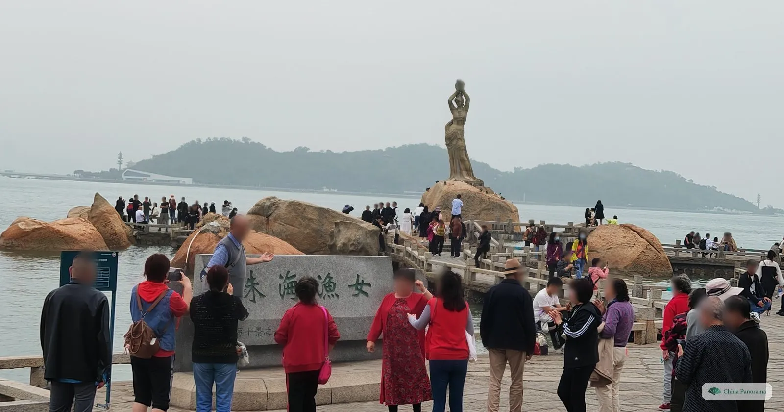 A granite statue of a woman holding a pearl above her head on a rock in the sea, a grey stone with the green characters 珠海渔女 in front and a crowd of visitors, an island behind