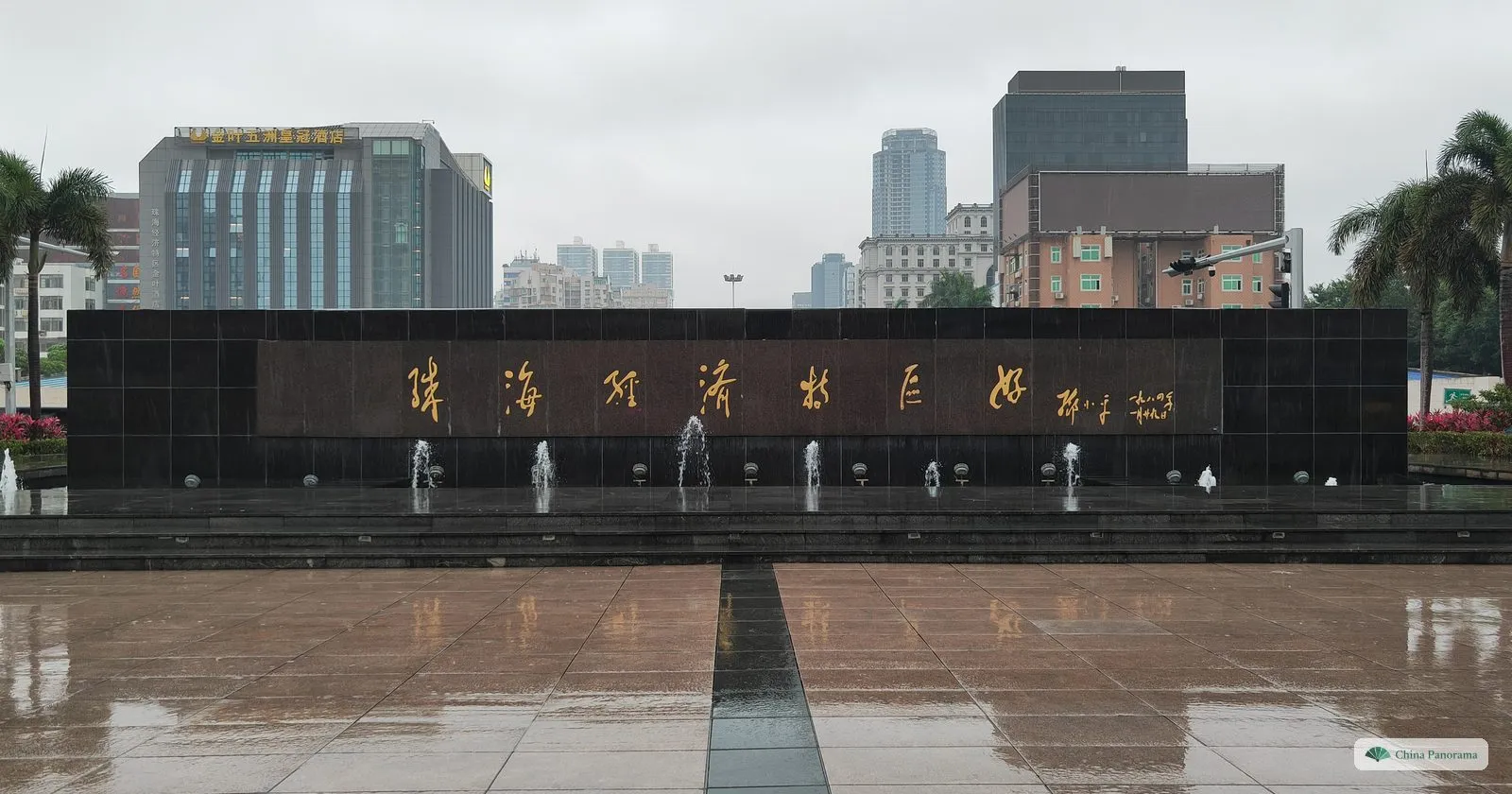 A long black stone wall with seven gold calligraphy characters, a signature and a date, low fountains in front and hotels and palms behind