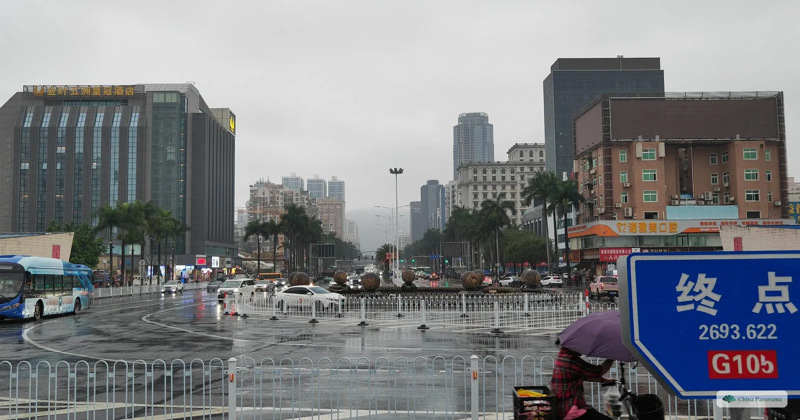 a wide wet road junction with a bus, a ring of bronze balls on a roundabout and a blue sign in the foreground reading 终点 2693.622 and G105