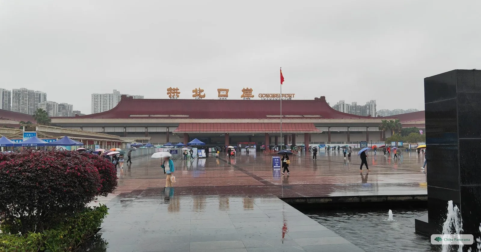 The same red-roofed border building seen from the side of the square, behind a black stone block with a fountain and a red-leaved hedge, in rain