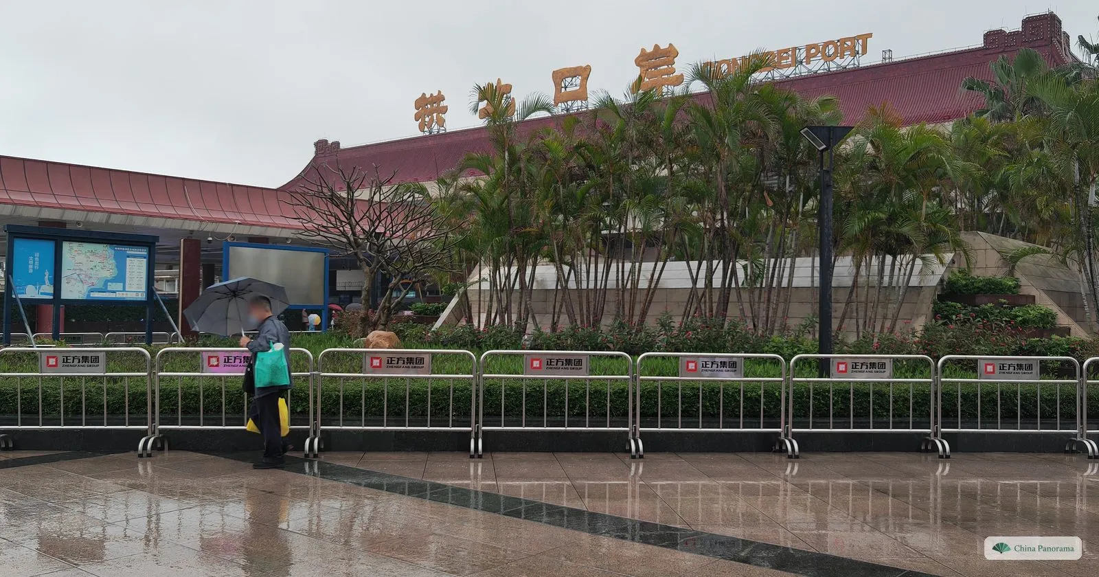 A red-roofed building behind a row of palms and metal barriers, with gold characters and “Gongbei Port” on the roof edge and a man with an umbrella by a map board