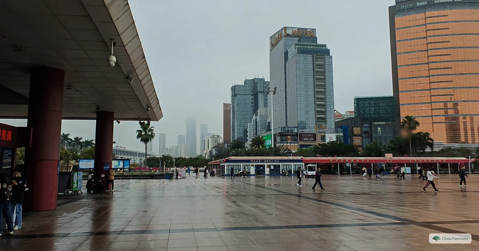 A wet square seen from under a canopy with red pillars, glass office towers and an orange-gold high-rise behind, people walking with umbrellas