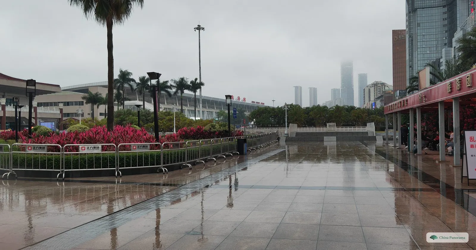 A wide wet paved walkway with a covered arcade on the right, beds of red-leaved plants behind barriers and tall towers in mist in the distance