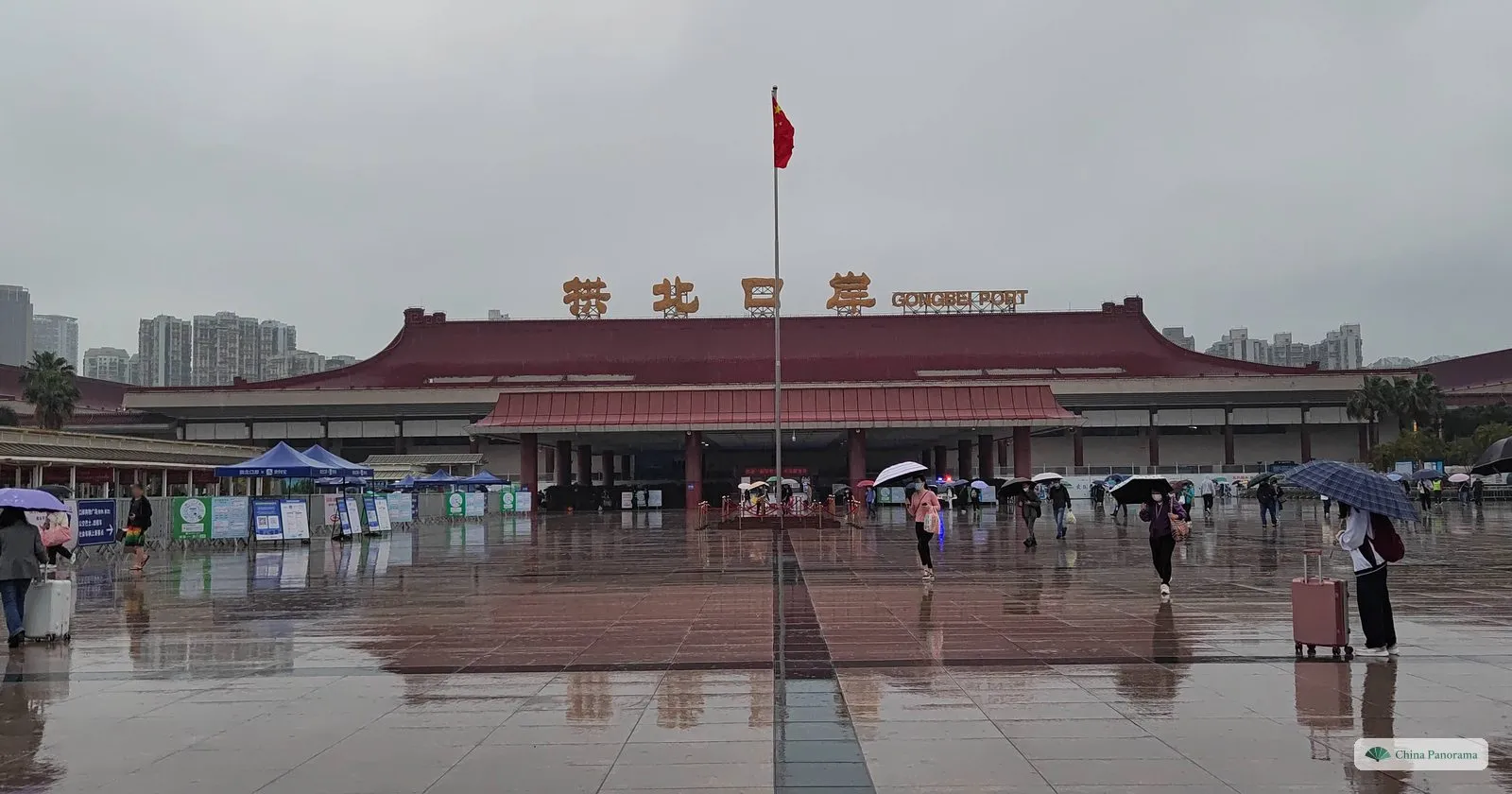 Gongbei Port: a long building with a red roof and gold characters, a flagpole and people with umbrellas on a wet square