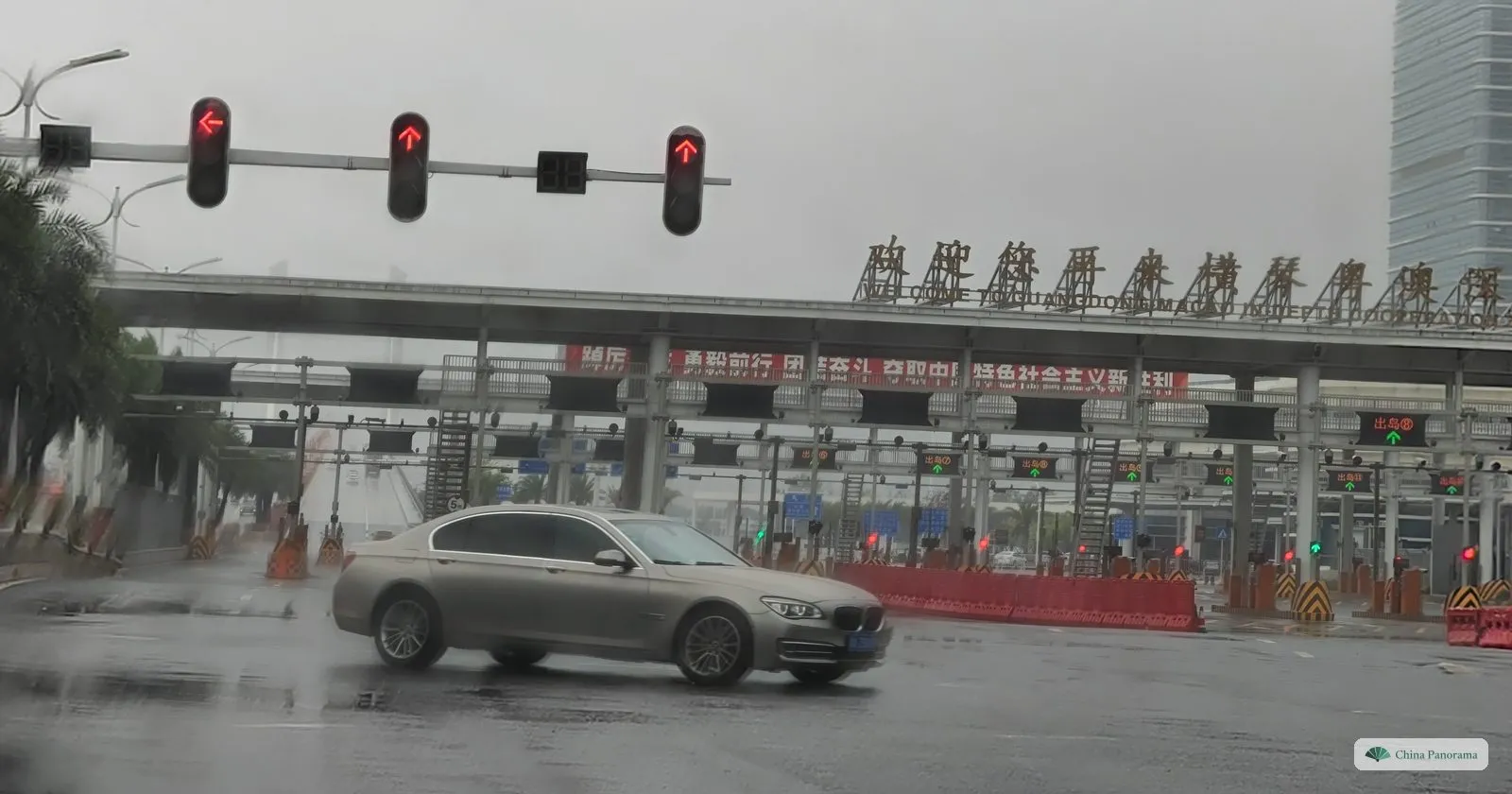 A row of toll-like gates under a long canopy with gold characters and red banners, green arrows over the lanes, a car turning in the rain