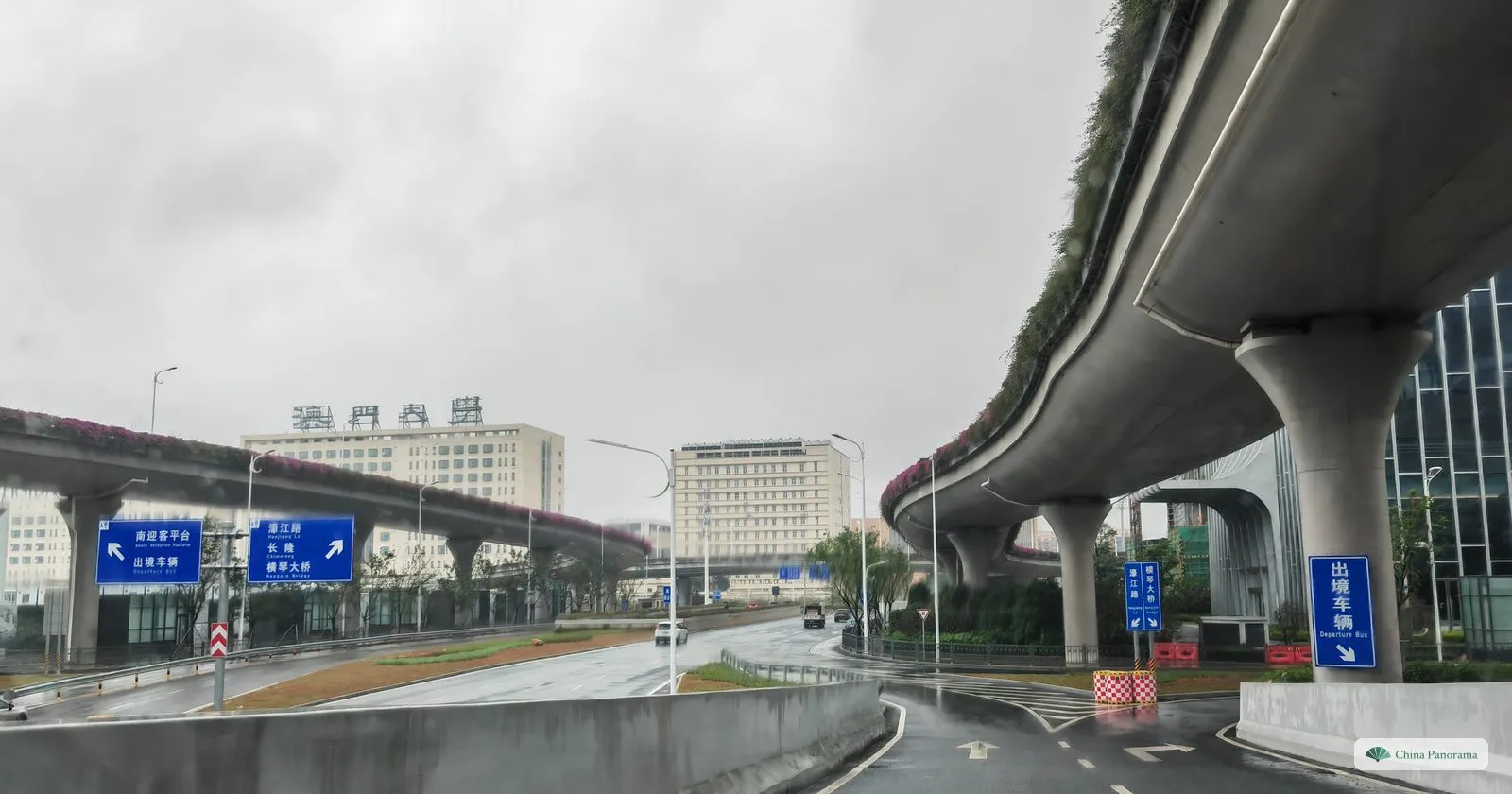 Elevated roads on round pillars with flowering planters, blue direction signs and a low white building with gold characters behind, on a wet day