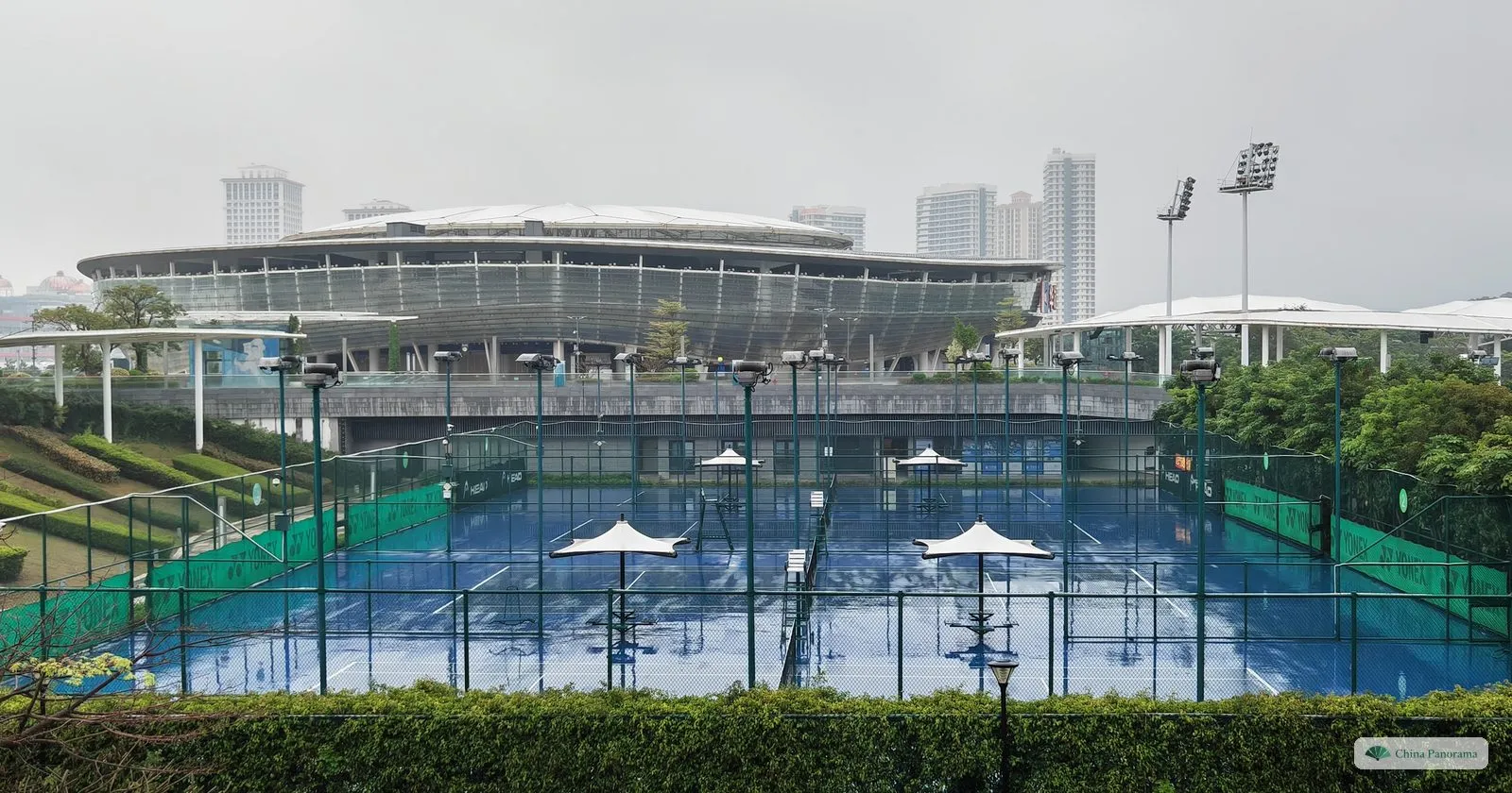 A tennis complex in the rain: blue hard courts with white shade umbrellas inside a fence, a stadium with a ring roof behind and towers in mist