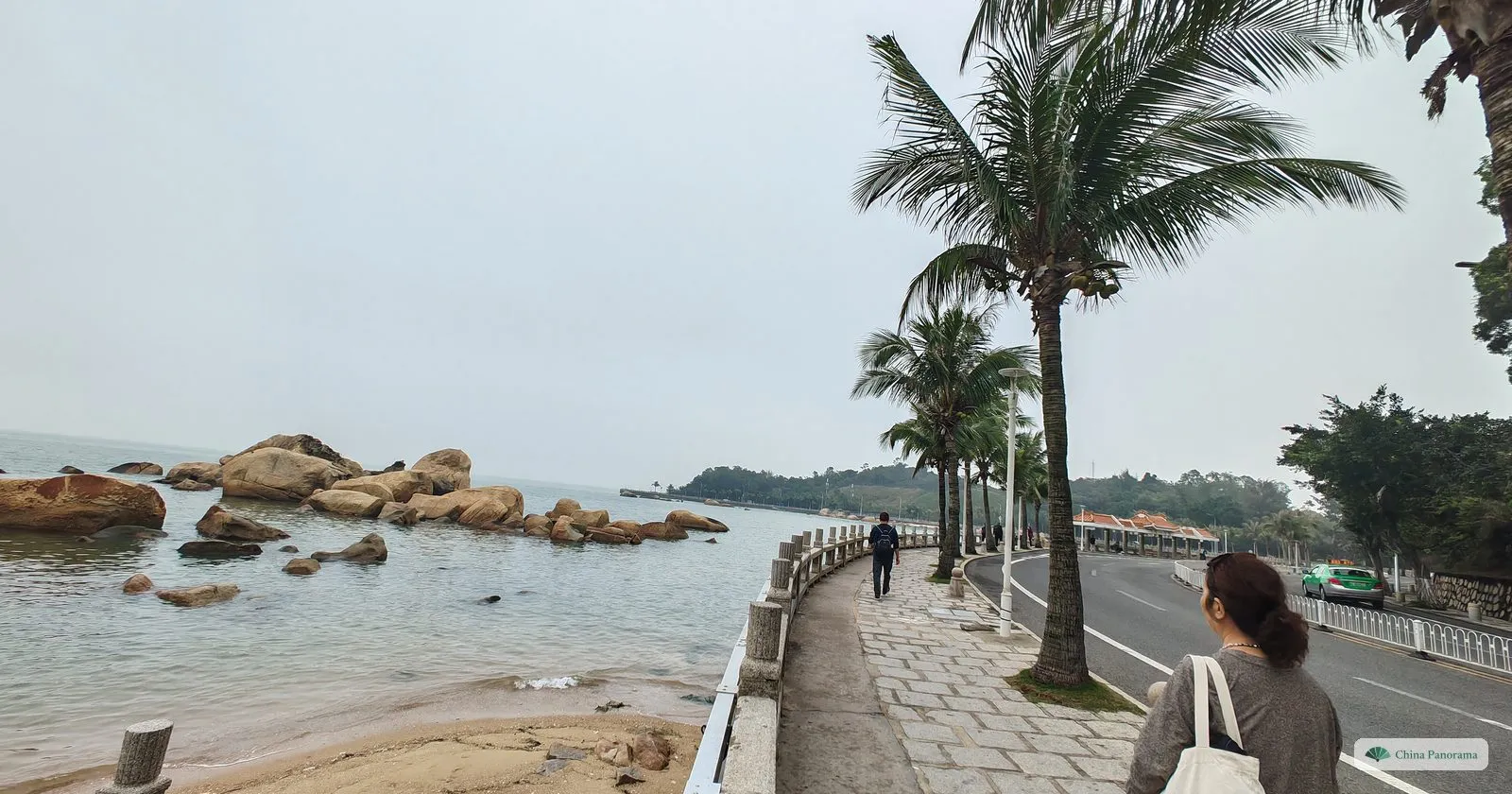 Rounded orange granite boulders in a calm grey bay beside a paved promenade with palms, a woman with a tote bag walking in the foreground