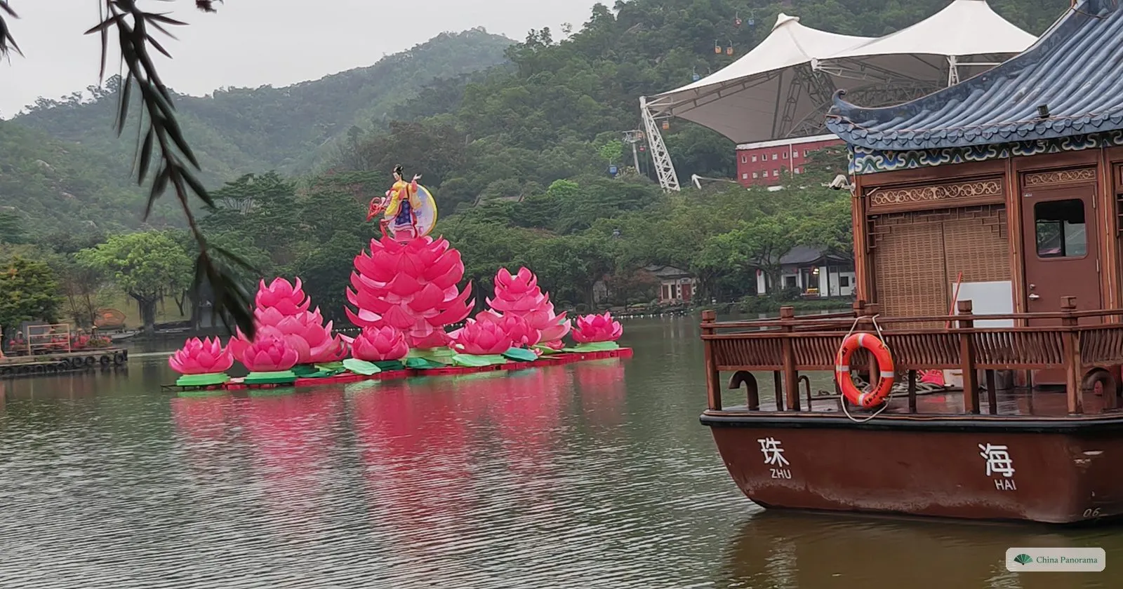 a wooden tourist boat with the characters 珠海 and “Zhu Hai” on its bow, pink lotus lanterns on the lake beyond and a tent roof on the hill