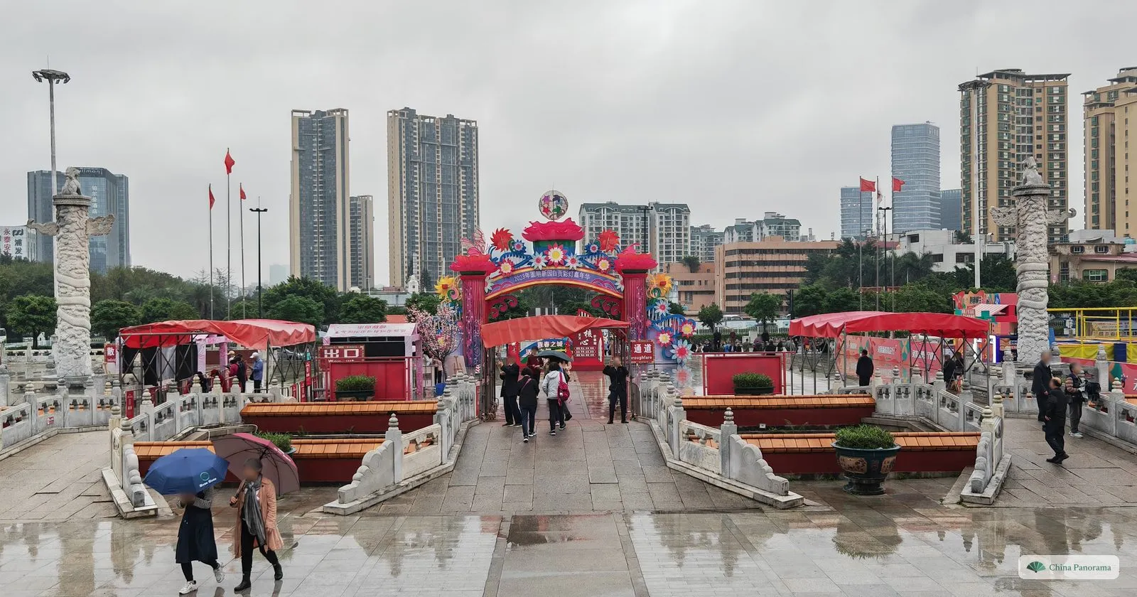 A plaza with white marble balustrades and two carved columns leading to a colourful lantern arch, people with umbrellas and high-rise flats behind