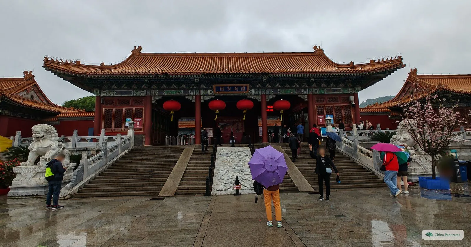 A Chinese palace gate with an orange glazed roof, red lanterns and a blue plaque, stone lions on either side of a staircase with a carved ramp, a visitor with a purple umbrella