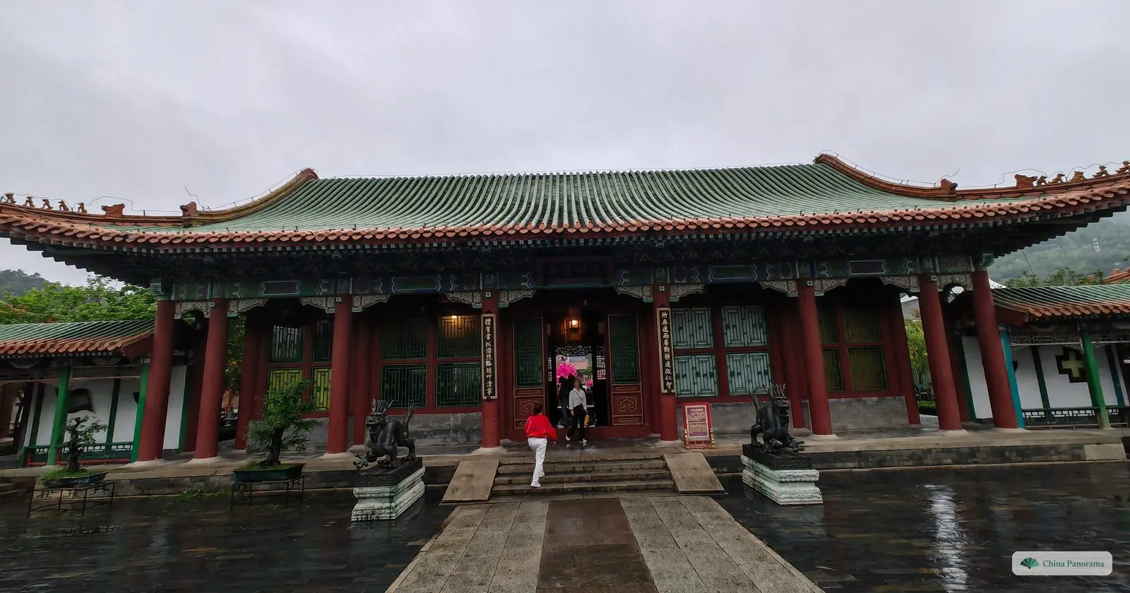 A side hall with a green tiled roof, red pillars and lattice windows at the end of a wet stone path, with bonsai trees in the courtyard