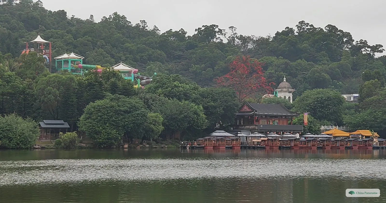 A lake with a wooded hill behind, a dark-roofed pavilion and a row of boat roofs on the far shore, a domed white building and a red-flowering tree among the trees