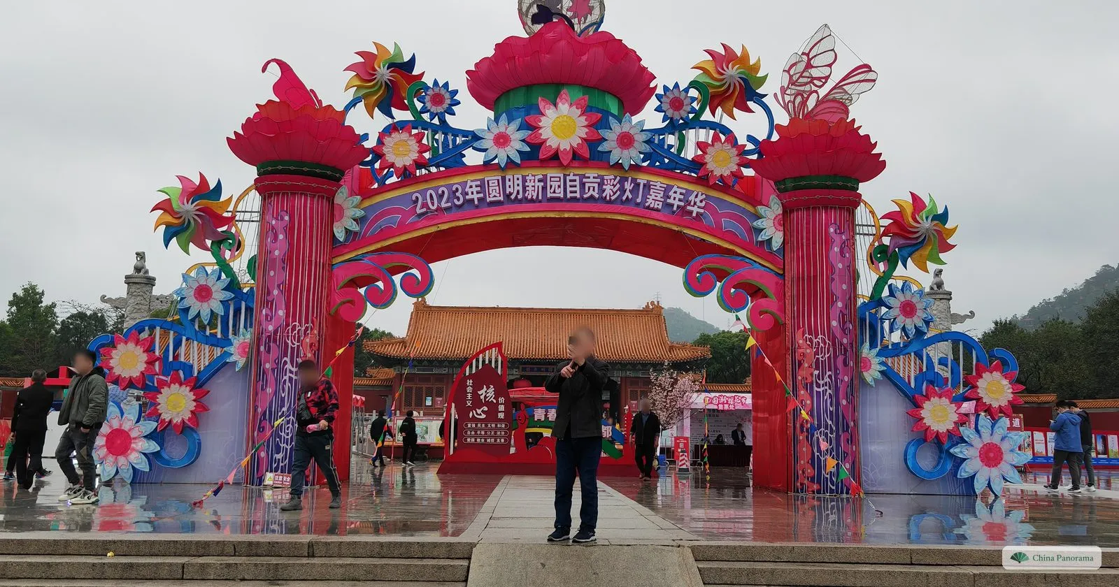 A huge arch of pink and blue lanterns shaped as flowers and pinwheels, reading “2023 New Yuanming Palace Zigong lantern carnival”, with a palace hall behind