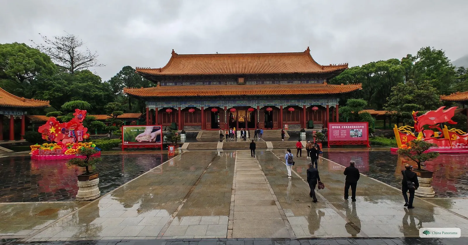 A wide wet square leading to a large two-storey hall with orange glazed roofs, flower displays and lantern figures on both sides