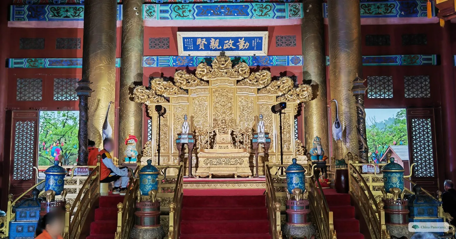 A gilded throne with carved dragons on a red-carpeted dais under a blue plaque with gold characters, golden pillars, white crane figures and blue enamel vessels on the stairs