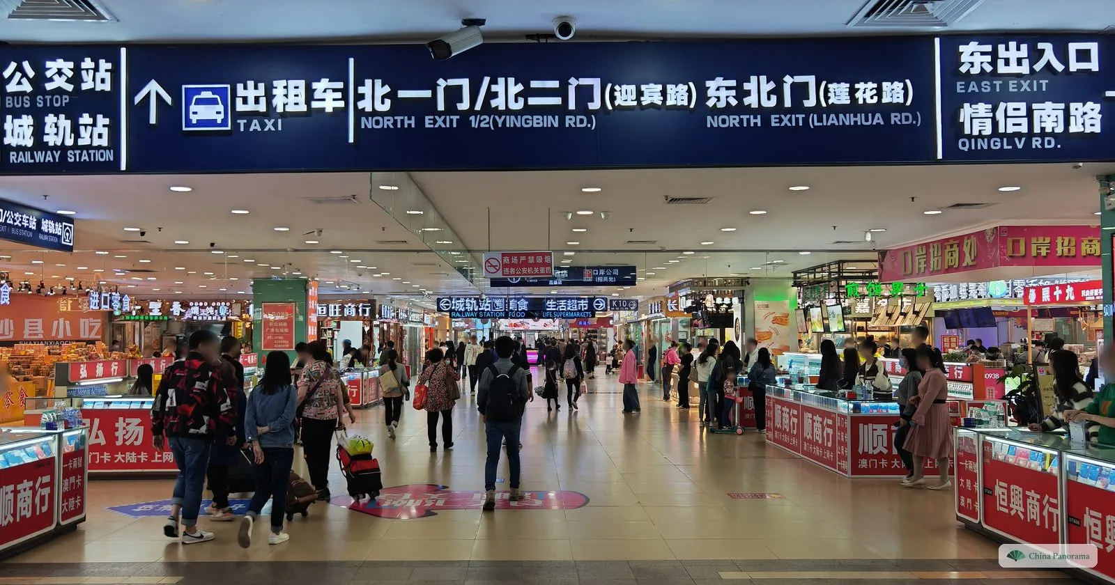 A long blue overhead sign in Chinese and English — bus stop, railway station, taxi, north exits, east exit to Qinglv Rd. — over a corridor of red counters
