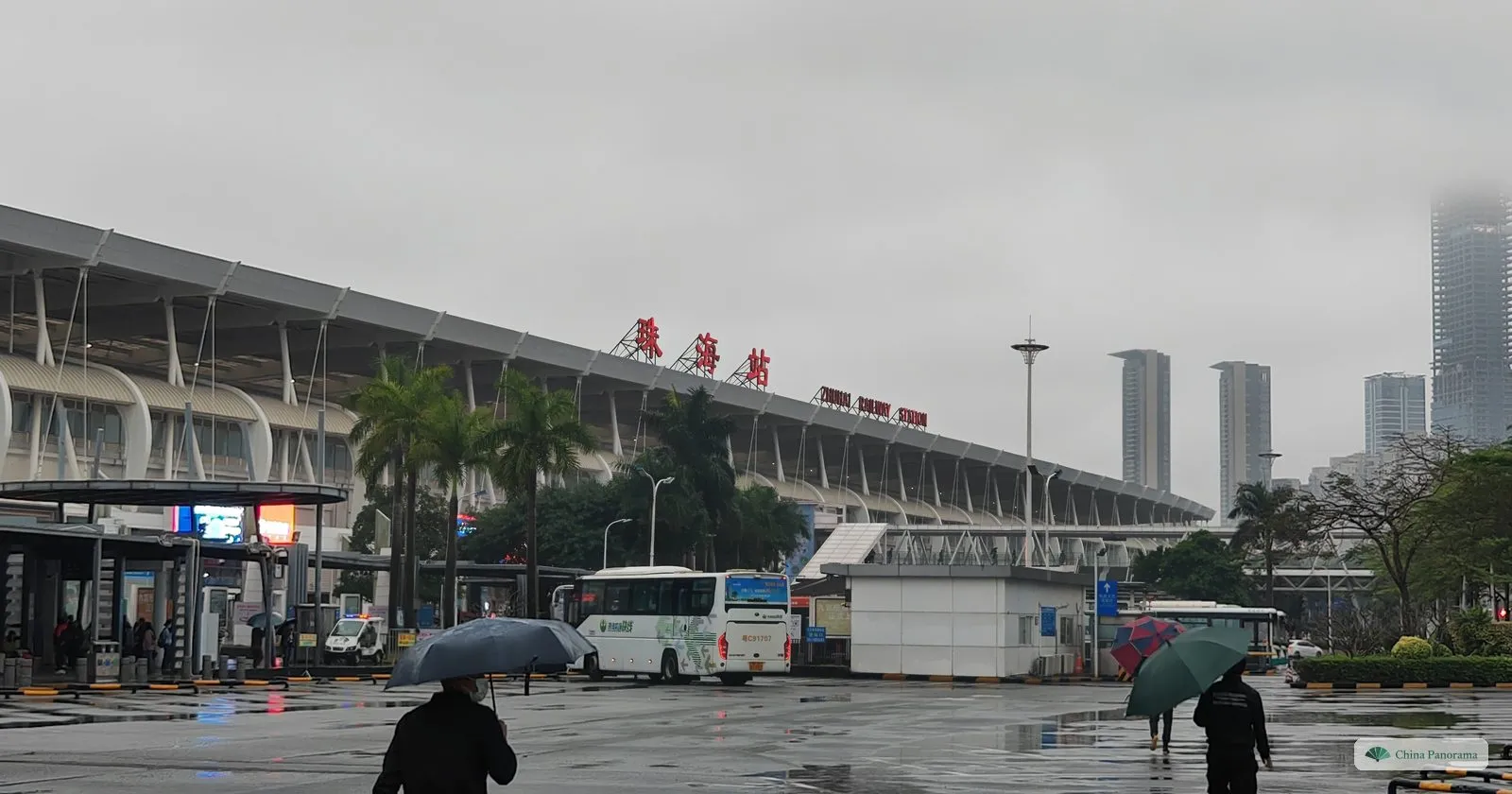 A long station building with a curved white roof and the red characters 珠海站, buses in front, two people with umbrellas on a wet forecourt